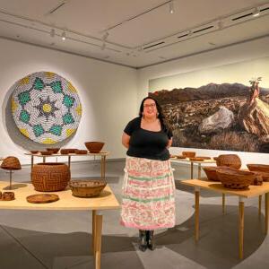 Meranda Roberts stands in the middle of a ring of Cahuilla baskets sitting on curved tables. In the background are installations of a basket made of cans and a scenic photograph of a desertscape.